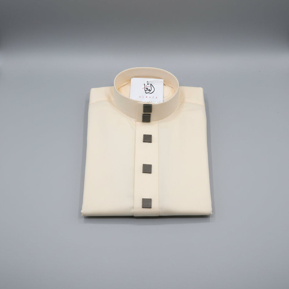 A folded cream-colored traditional thobe with a mandarin collar and four dark square buttons, displayed on a wooden surface with a brand tag inside the collar.