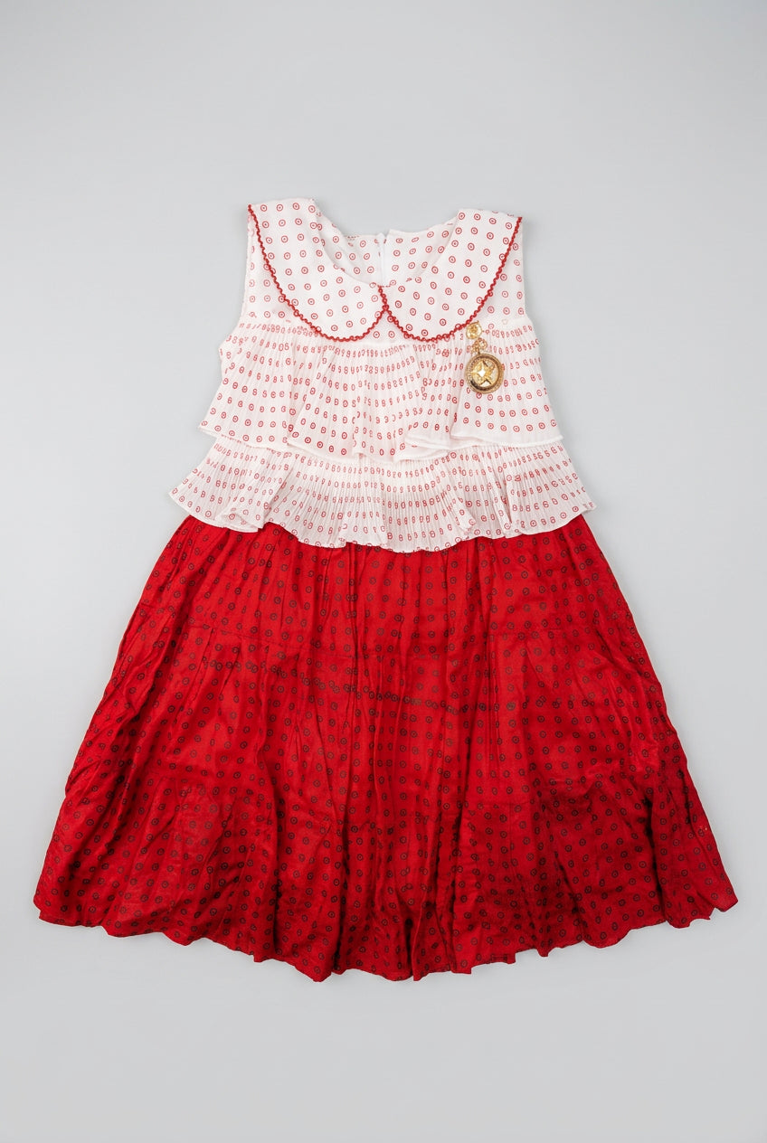 A sleeveless children's dress laid flat on a cardboard box. The top is white with a red polka dot pattern and a ruffled Peter Pan collar, while the skirt is a solid red with a small black pattern.