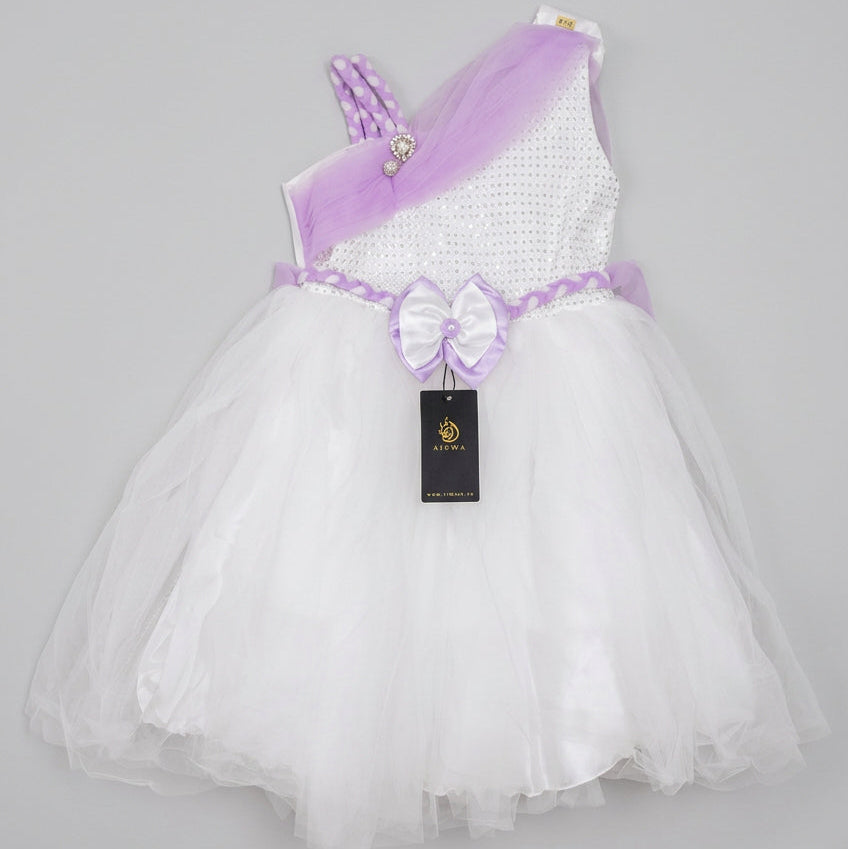 A toddler girl's one-shoulder tulle dress with a white textured bodice, lavender purple sash, and matching bow on a white tulle skirt, laid flat on a brown cardboard box.