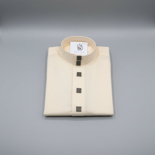 A folded cream-colored traditional thobe with a mandarin collar and four dark square buttons, displayed on a wooden surface with a brand tag inside the collar.