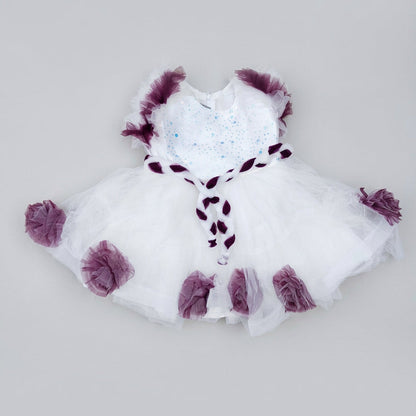 A white tutu-style dress with deep purple ruffled trim and a matching purple ribbon detail, laid flat on a large piece of brown cardboard.
