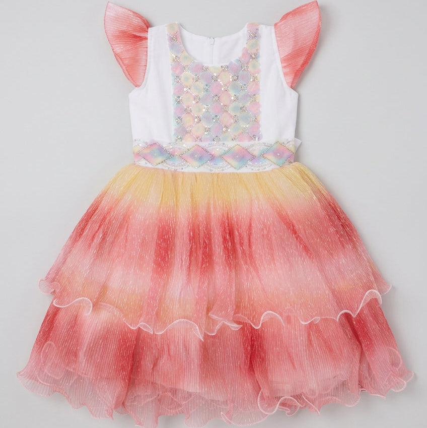 A children's party dress laid flat on a cardboard box. The dress has a white bodice with a pastel mermaid-scale pattern, pink ruffled shoulder straps, and a tiered tulle skirt in a pink, orange, and yellow ombre gradient.