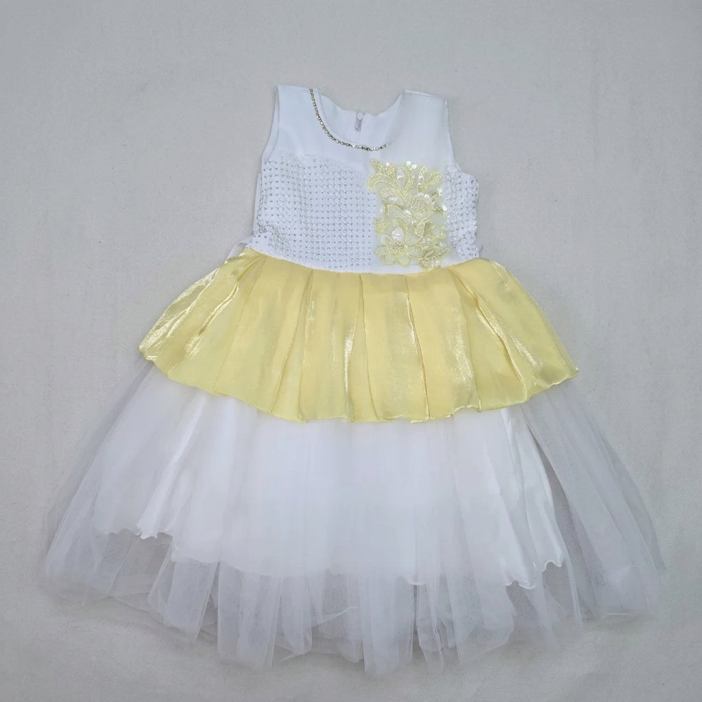 A children's party dress laid flat on a brown cardboard sheet, featuring a white sleeveless bodice with a yellow floral applique, a yellow ruffled skirt, and a layered white tulle tutu.