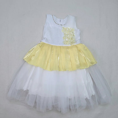 A children's party dress laid flat on a brown cardboard sheet, featuring a white sleeveless bodice with a yellow floral applique, a yellow ruffled skirt, and a layered white tulle tutu.