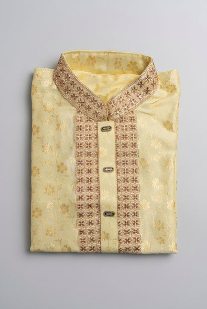 A folded light yellow kurta with gold floral patterns and brown embroidered trim on the collar, placket, and front buttons, displayed on a dark textured background.